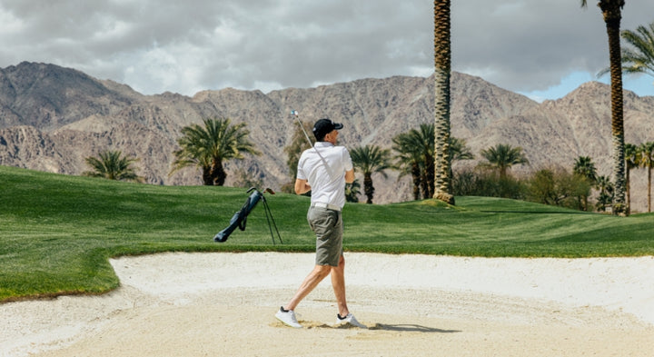 the follow through of man in shorts and wearing hat hit a golf ball from a fairway sand trap with a golf bag, palm trees and mountains in the background