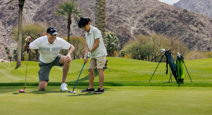 man kneeling down on a green with putter in hand behind a young junior putting on a putting green with two golf bags, palm trees and mountain in the background