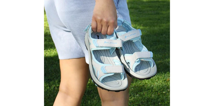 woman with shorts carrying pair of light gray/aqua Orlimar Ladies Golf Sandals with Replaceable Spikes