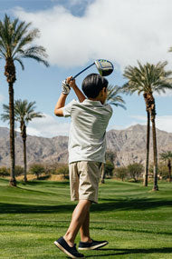 follow through of a young kid hitting an Orlimar junior driver down the fairway with palm trees and mountain in the background