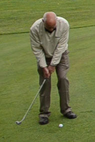balding man wearing a pullover jacket getting ready to hit a chipper golf club from off the green