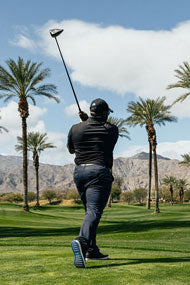follow through of a male golfer teeing off on a golf course with palm trees and mountains in the background