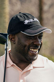 smiling man with a golf club resting on his shoulder wearing a black Orlimar golf hat