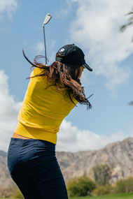 follow through of a woman golfer wearing a yellow shirt and hair flowing beneath a black golf hat hitting an iron