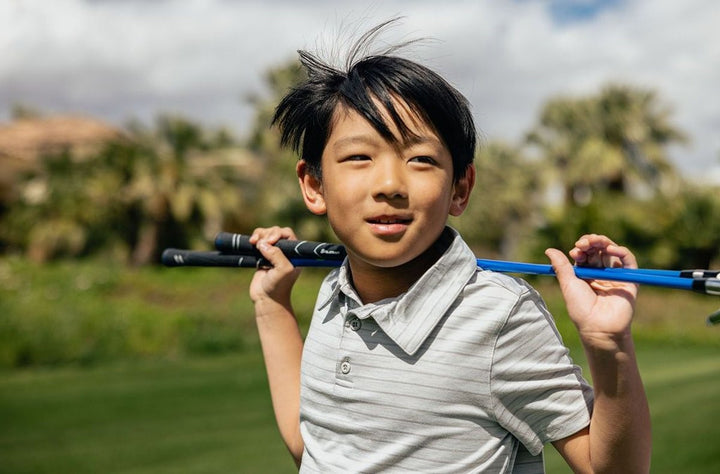 Young Asian boy smiling and holding two Orlimar junior golf clubs horizontally against his back