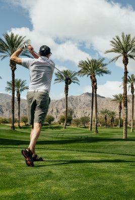 follow through of man wearing a hat swinging an Orlimar golf driver off a tee box surrounded by palm trees and mountain on a partly cloudy day 