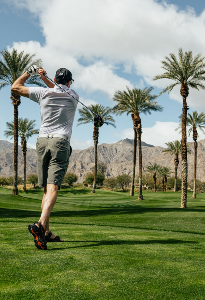the follow through of a man with a hat swinging an Orlimar golf driver on a tee box with palm trees and mountain in the background 