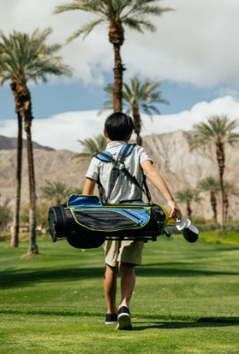 young kid carrying a black and blue Orlimar golf bag with double carry straps while walking a golf course with palm trees and mountain in the background