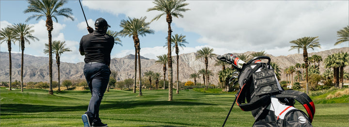 golfer teeing off on a golf course with palm trees with a black golf bag off to the side of the tee box