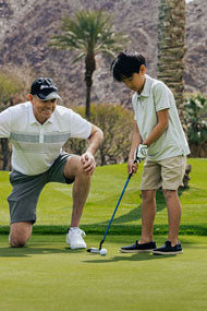 kneeling man behind a young junior golfer ready to putt on the green